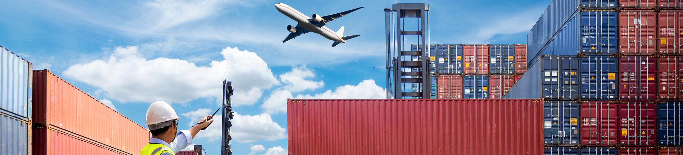 Pane flying over shipping containers with man pointing to the sky in hard hat.